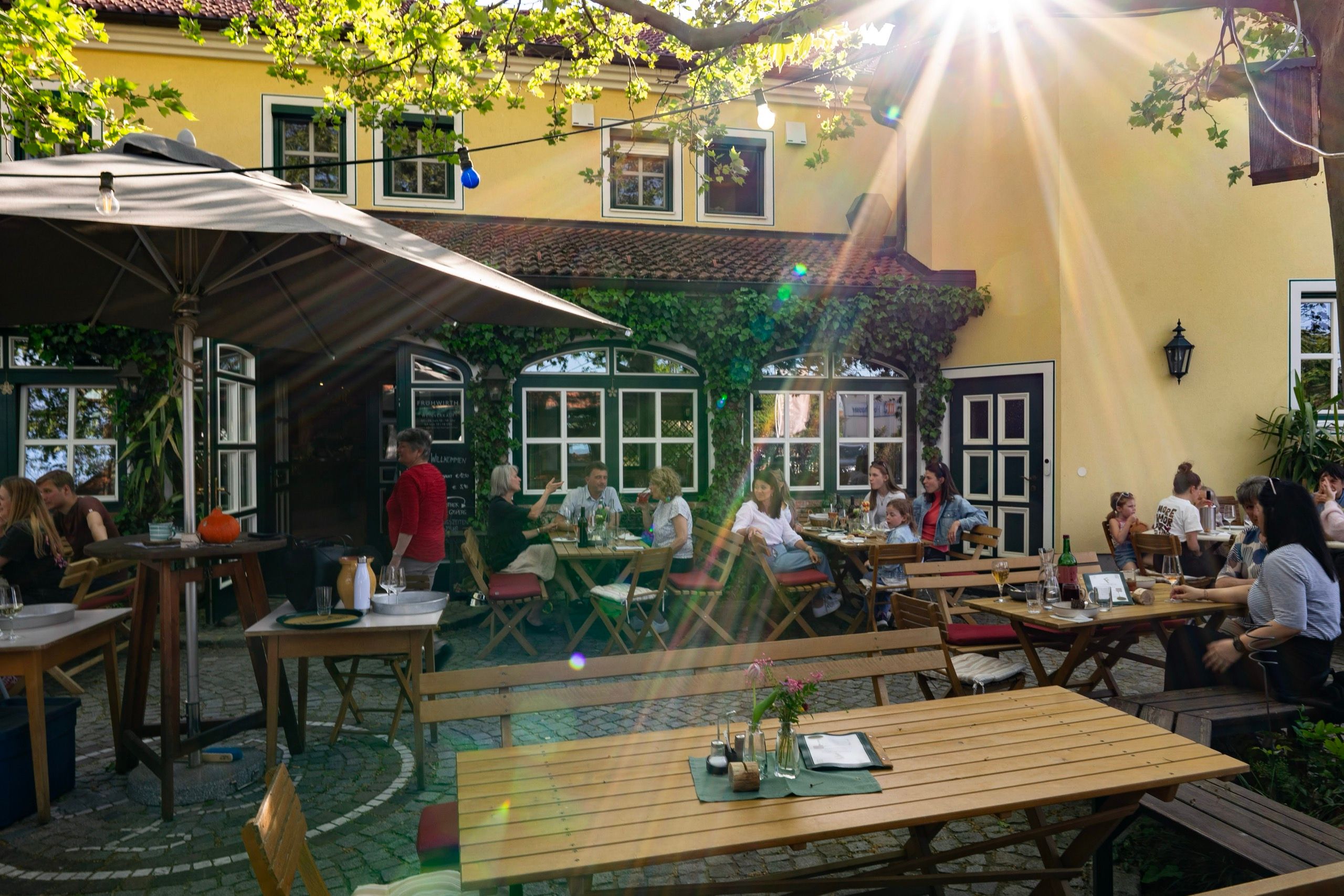 Gastgarten des Heurigen Frühwirth im Gegenlicht: gedeckte Tische unter Bäumen, Gäste in der Nachmittagssonne