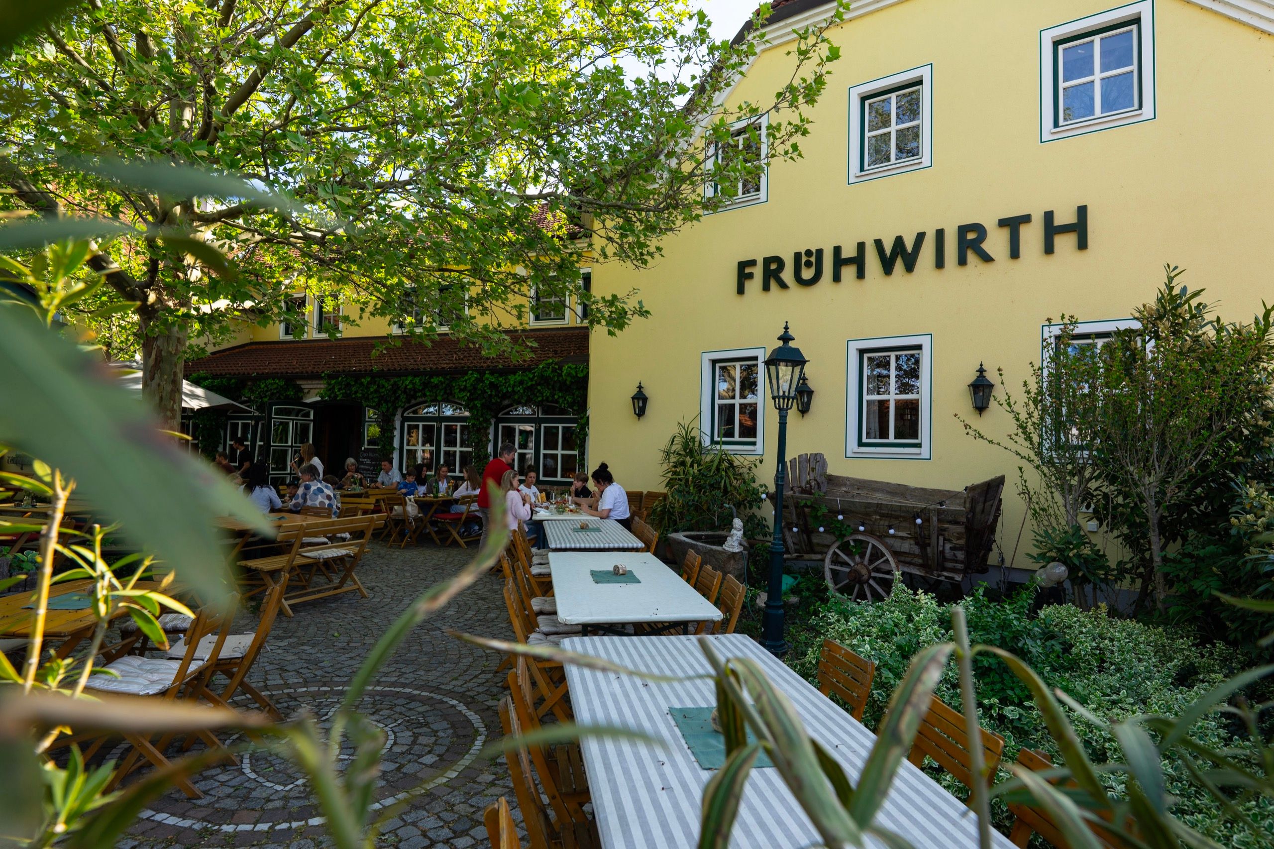 Das gelbe Haupthaus des Weinguts Frühwirth mit Schriftzug an der Fassade, davor der Gastgarten mit langen Tischen unter Bäumen.