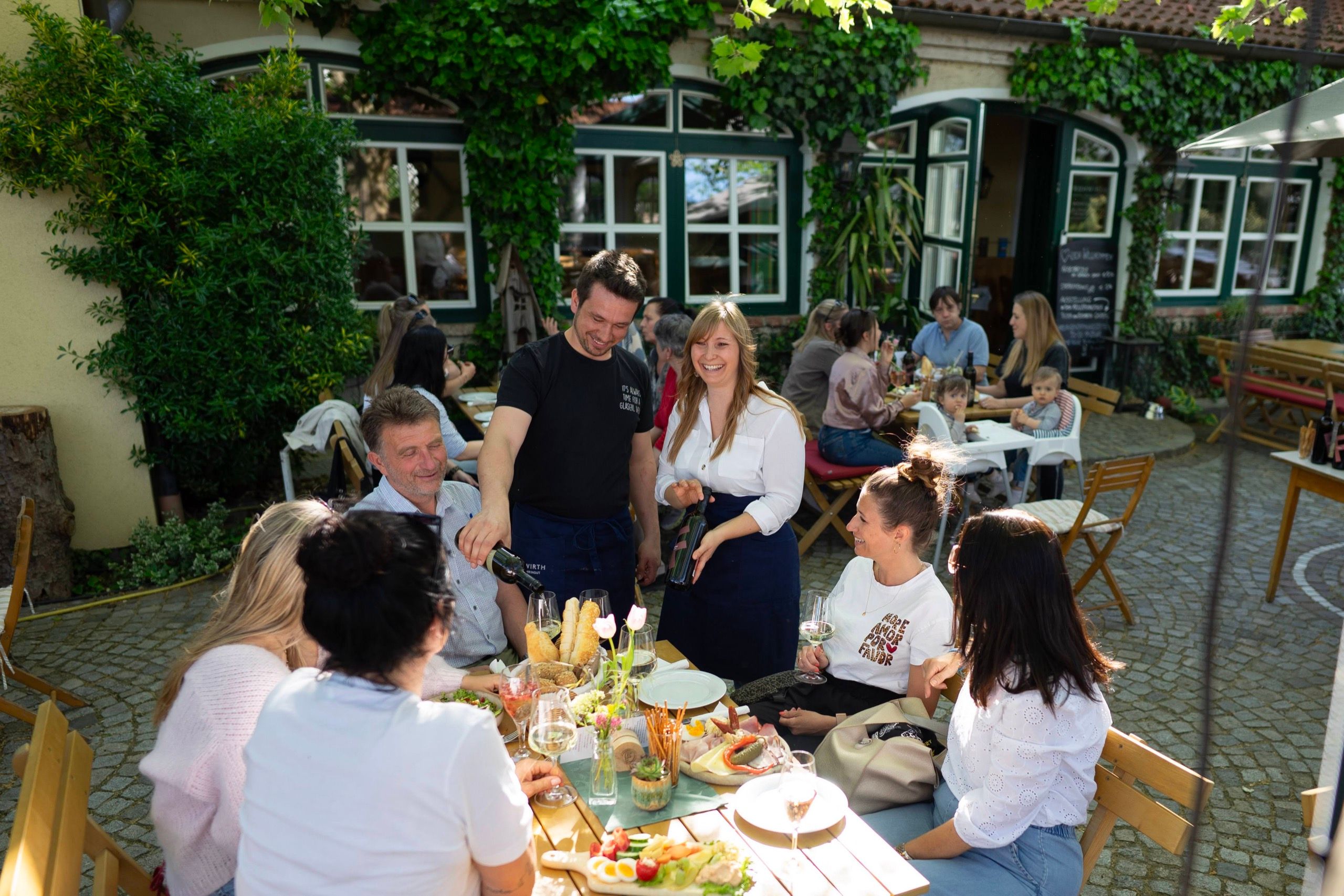 Im Gastgarten des Heurigen: Service schenkt Wein aus, Gäste an einem langen Tisch mit Brettljause