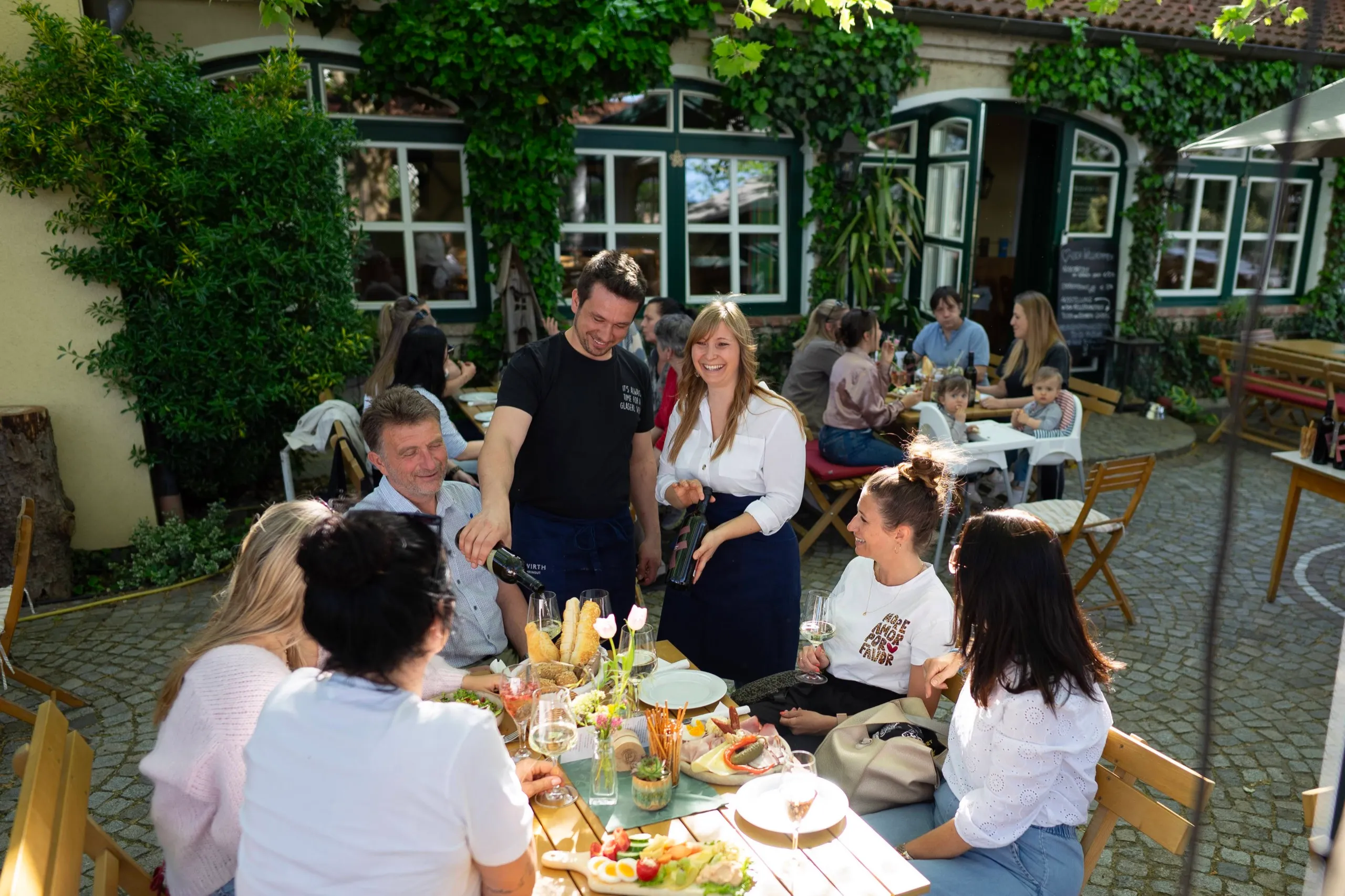 Service schenkt Wein ein an einer gedeckten Tafel im Gastgarten, Gäste ringsum, dahinter das Heurigenhaus