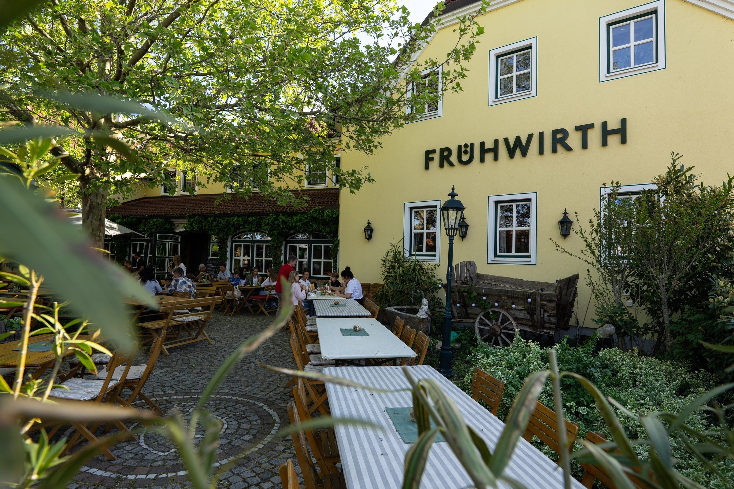 Das gelbe Haupthaus mit FRÜHWIRTH-Schriftzug, davor der Gastgarten unter Bäumen
