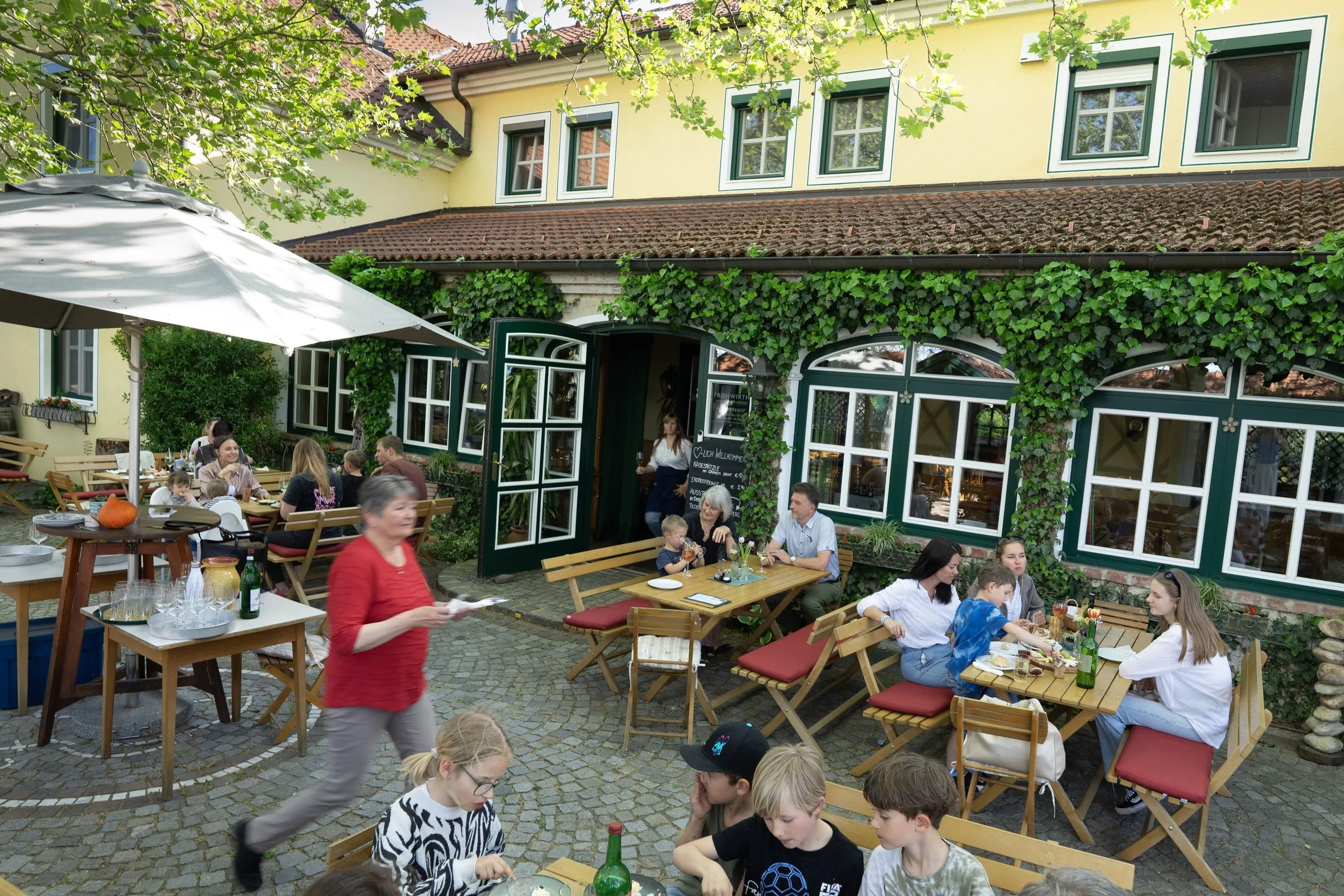 Gastgarten mit Tischen unter alten Platanen, Gäste, im Hintergrund das gelbe Haupthaus