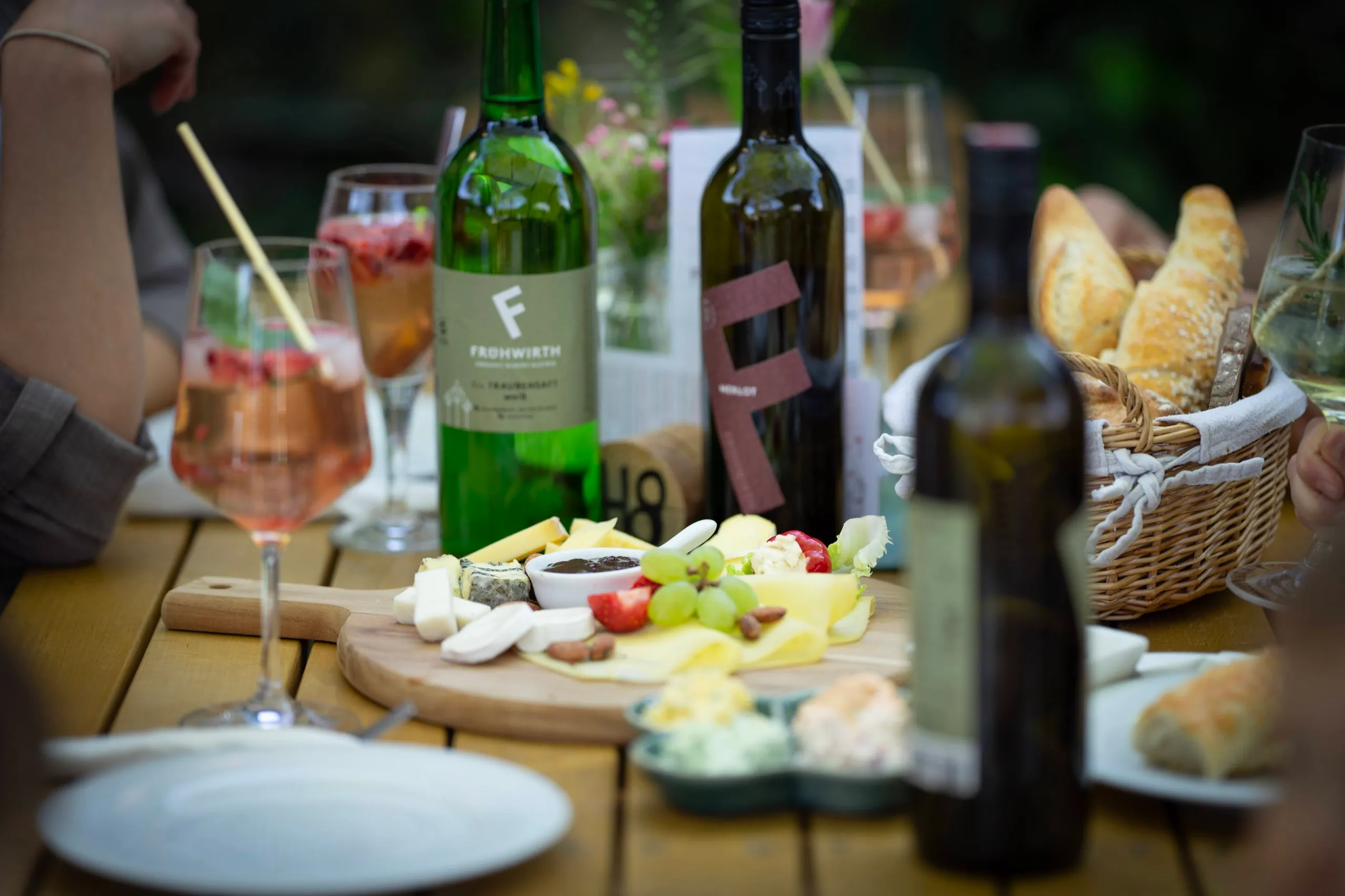 Gedeckter Holztisch im Gastgarten: Jausenbrett mit Käse, Aufstrich und Trauben, dazu Brotkorb, Weinflaschen und ein Glas Spritzer